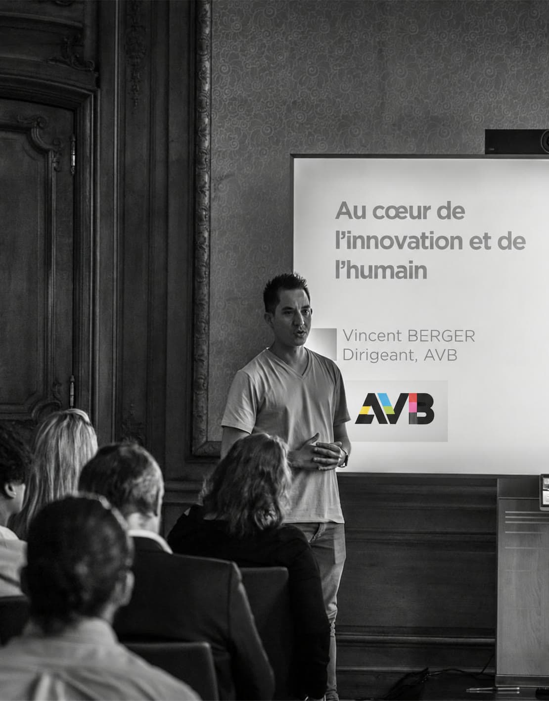 Vincent Berger parle debout face au public, devant l'écran de sa présentation au logo AVB, dans un salon aux boiseries anciennes