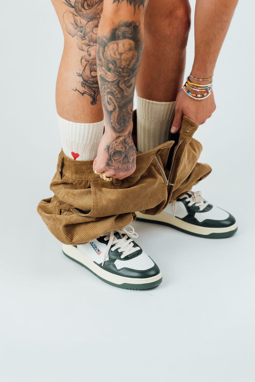 Shooting mode Contresens : un homme tatoué enfile un pantalon en velours côtelé camel, chaussettes écrues et baskets Autry