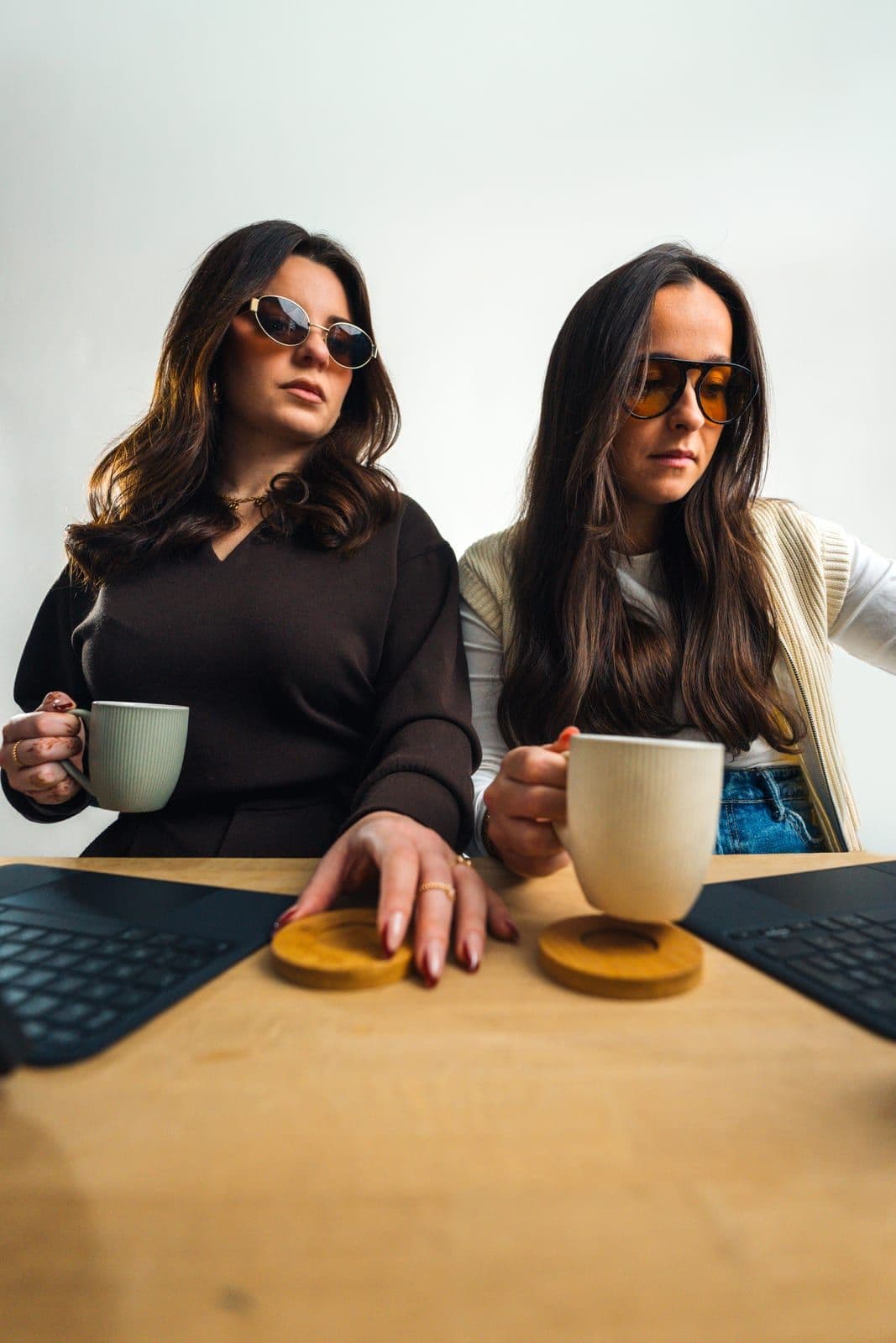 Deux femmes en lunettes de soleil, une tasse de café à la main, derrière une table et des claviers